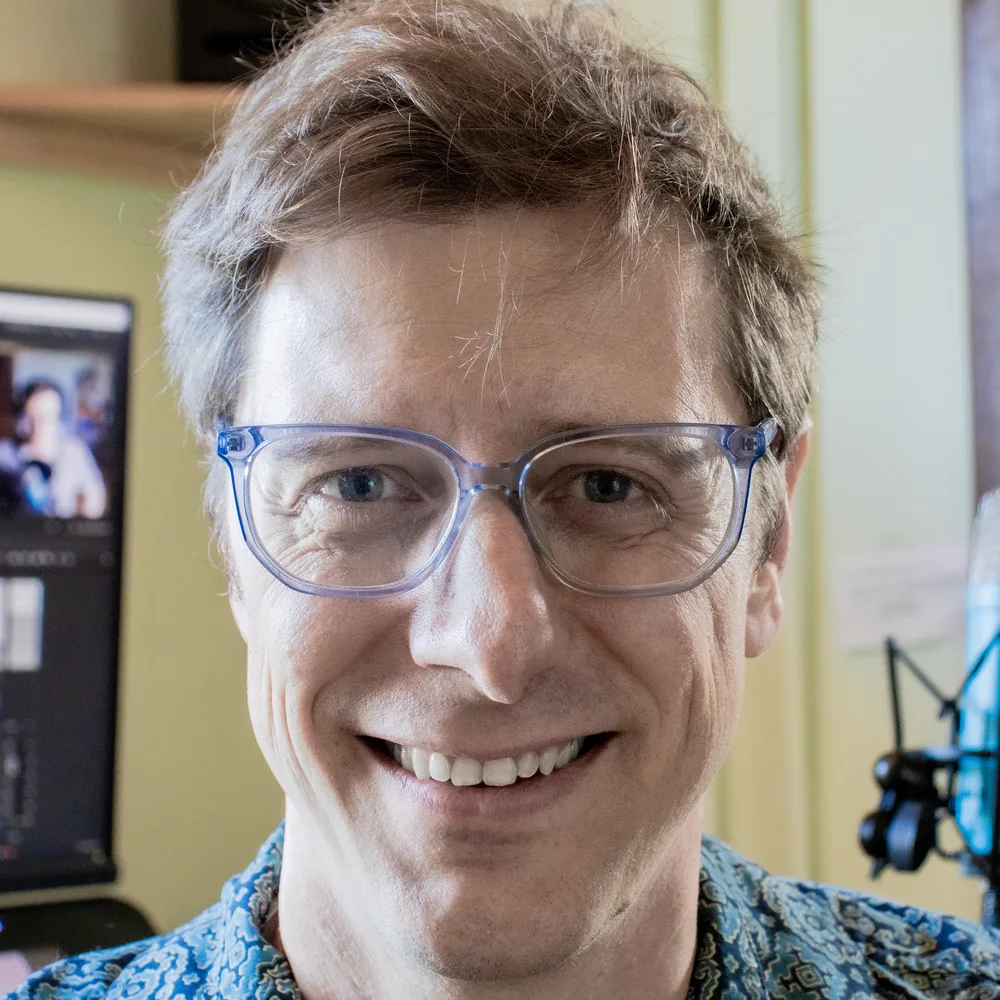 Charlie LaVoy, smiling, in clear-framed glasses and a patterned shirt, in his studio